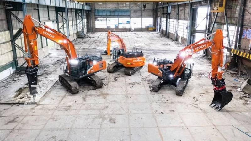 Four orange Hitachi excavators parked inside a large, empty industrial bay with a yellow overhead crane above.