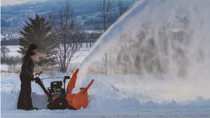 Person operating an orange snow blower, clearing snow in a snowy yard with bare trees in the background.