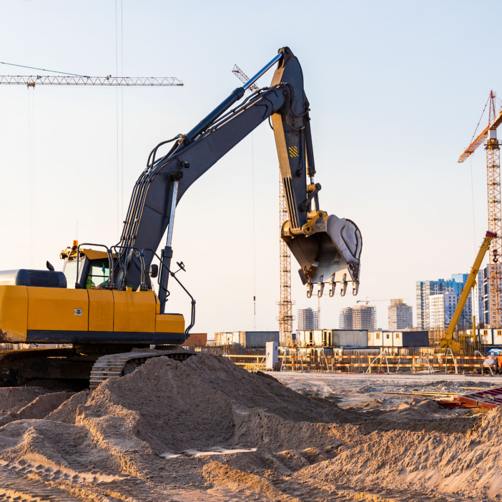 Yellow excavator with extended arm and bucket at a construction site; cranes and buildings in the background.