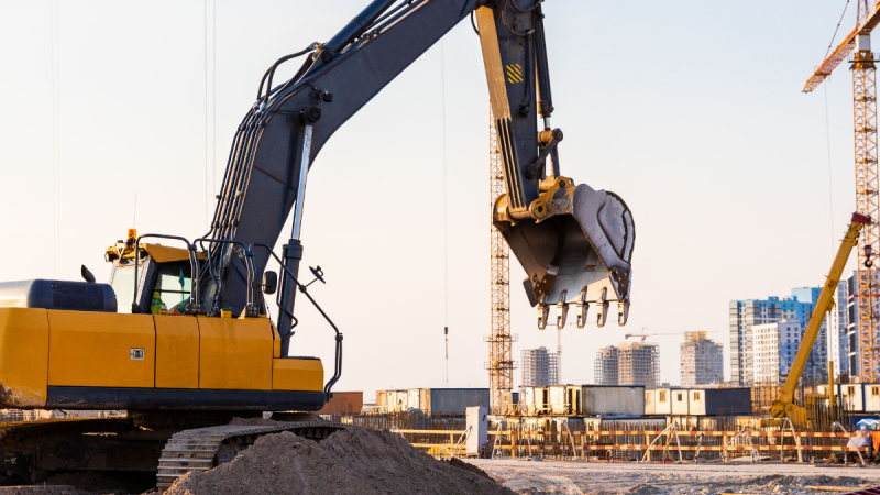 Yellow excavator with extended arm and bucket at a construction site; cranes and buildings in the background.