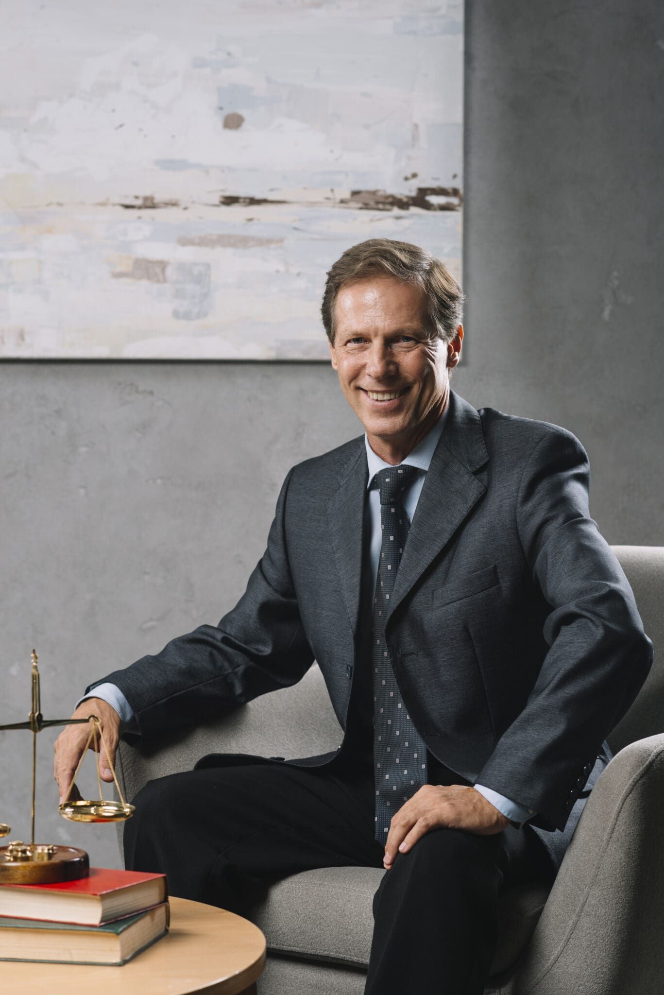 Smiling man in a suit sitting in an office with law books and a scale on the table.