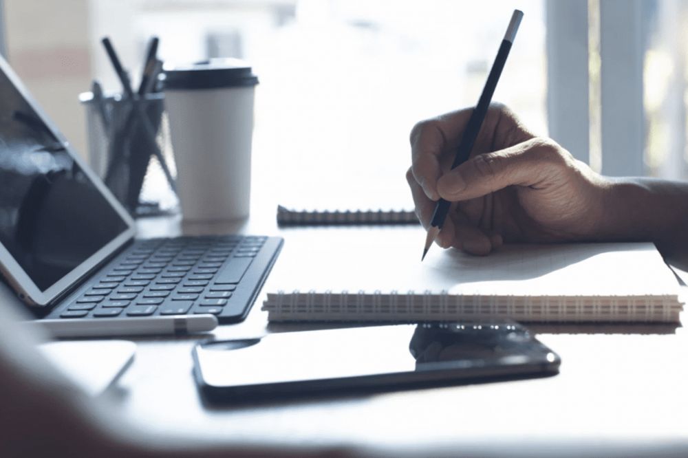 Hand writing notes at a desk with a tablet, smartphone, and coffee, reflecting a productive workspace.