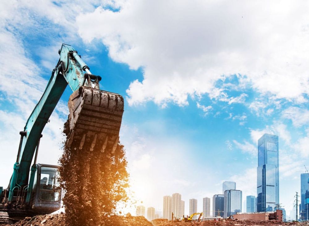 Excavator at construction site with city skyline in background under clear blue sky.