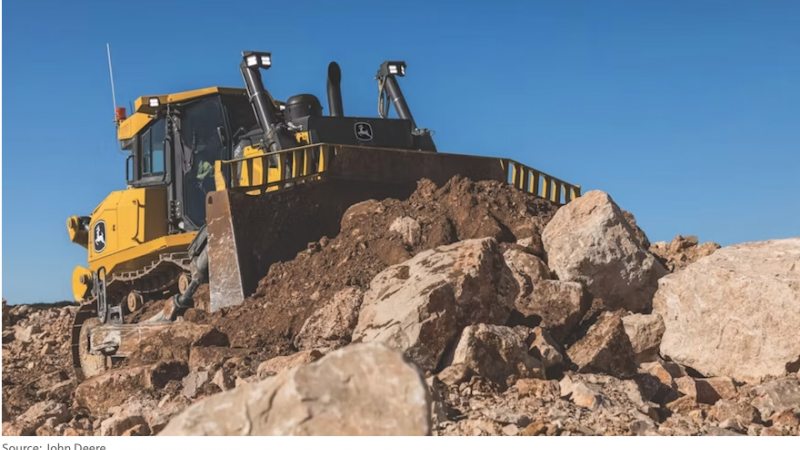Yellow bulldozer with a large blade on a pile of dirt and rocks at a construction site against a blue sky.