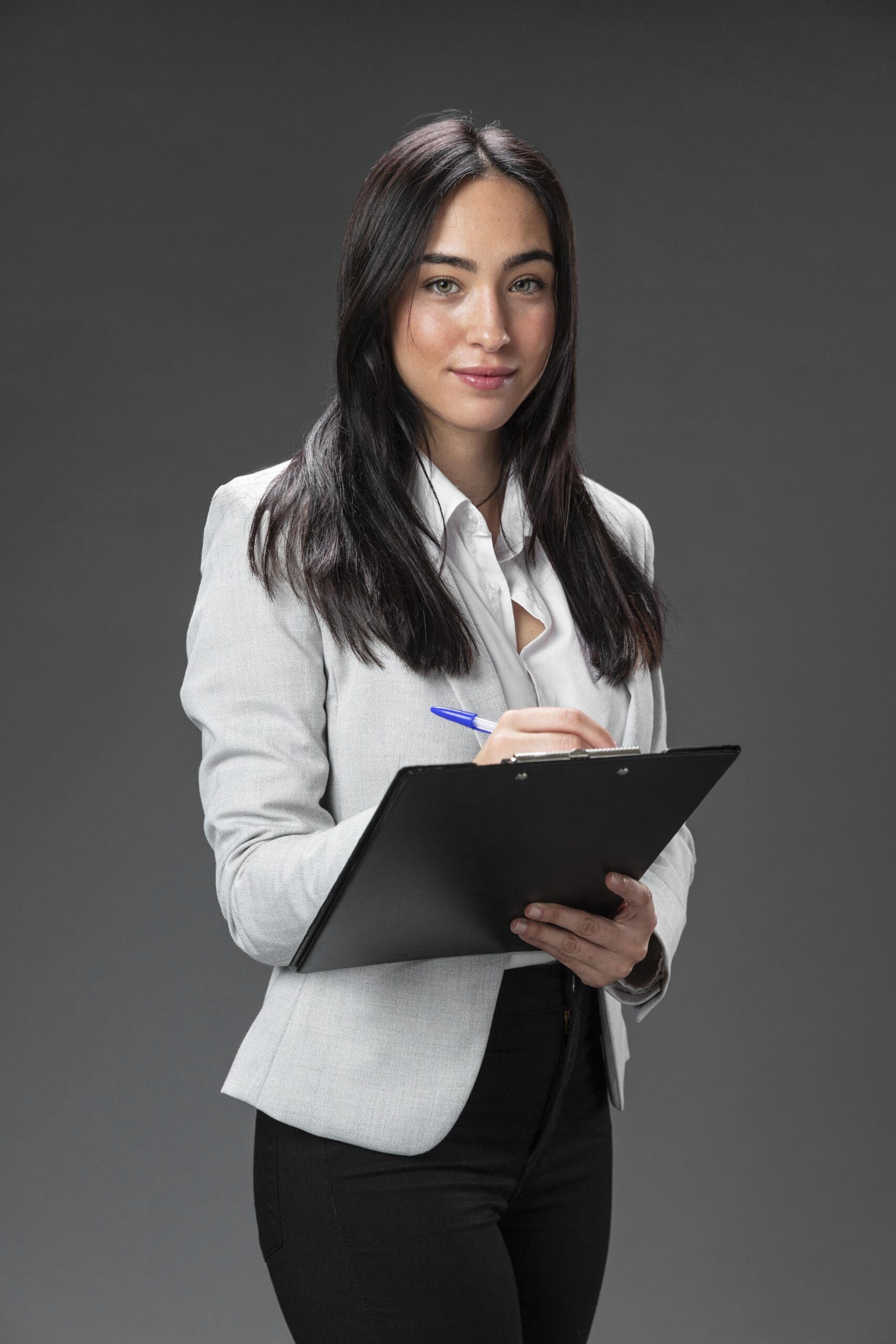 Professional woman in business attire holding a clipboard, confident and poised against a gray background.